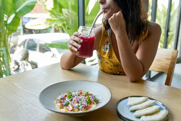 Unposed, natural shot of beautiful brunette vegan businesswoman in a fashionable yellow floral dress eating tasty fine, organic, international cuisine on artisanal ceramic plate including naan bread, tomatoes and herbs drinking a red vegetable juice from a reusable metal straw.