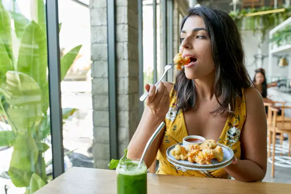 Unposed, natural shot of pretty brunette ethnic vegan model eating tasty fine vegetarian plant-based cuisine on artisanal ceramic plate including fried cauliflower and sauce with a green smoothie and reusable metal straw for mindful eating and a nutritious paleo diet.