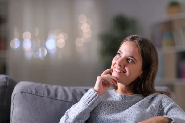Happy woman thinking looking at side sitting on a couch in the living room in the night at home