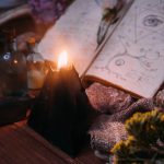 Open old book with magic spells, runes, black candle and herbs on witch table.