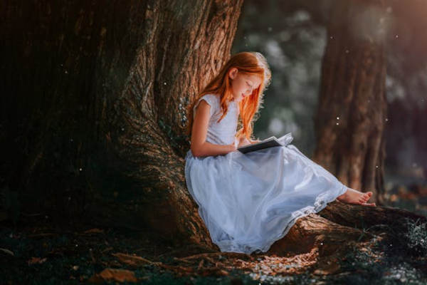 young girl in white dress leaning on a tree trunk holding and reading a book